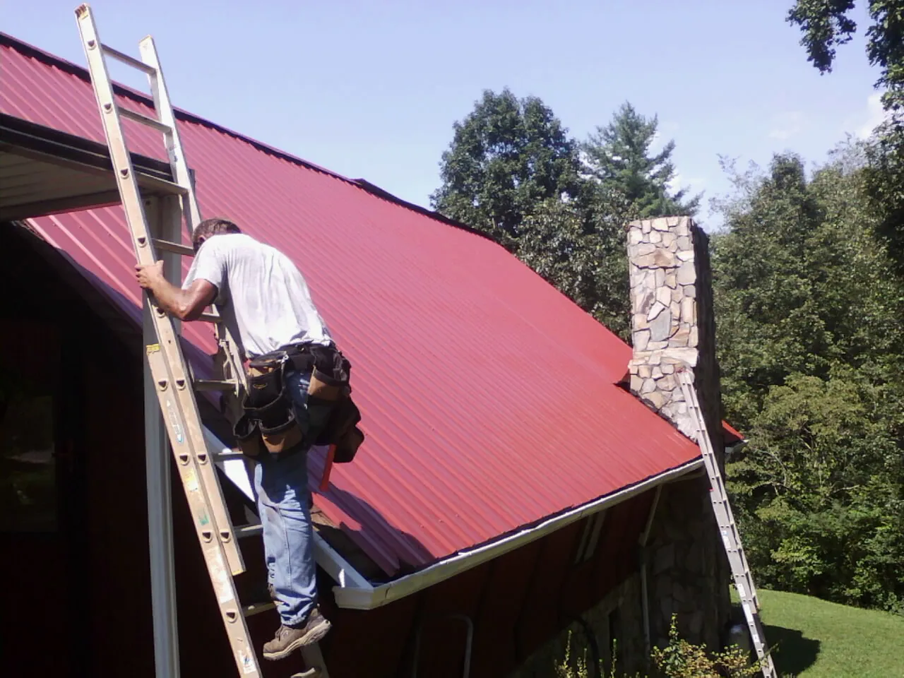 Roofer on a ladder beside a red metal roof and stone chimney — Soffit & Fascia Repair by Roofing Brothers in Wawarsing